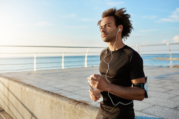 Jogger masculin élégant avec une coiffure touffue à la recherche directe le matin, profitant d'activités sportives. Mettre l'homme dans les écouteurs avec une bouteille d'eau dans les mains en prenant une pause au milieu de la formation