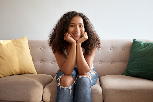 Joie, bonheur, loisirs et émotions positives. Jolie fille à la peau sombre dans des vêtements élégants se détendre dans le salon sur un canapé confortable, tenant les mains sur son menton et souriant joyeusement