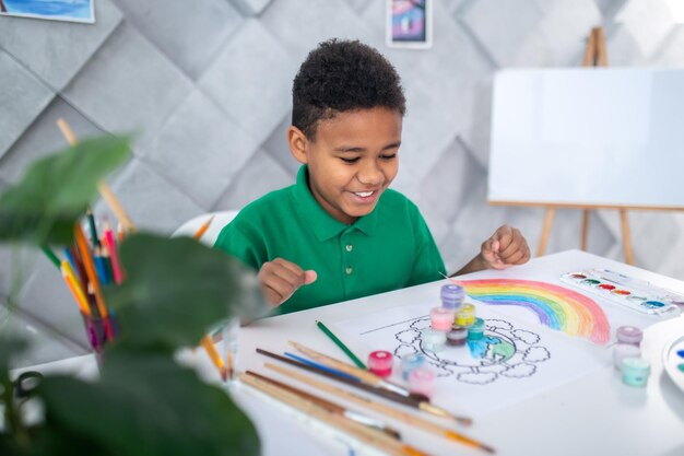 Joie. Heureux garçon à la peau foncée d'âge scolaire dans des vêtements décontractés assis à table regardant joyeusement la pyramide construite de peintures dans une salle lumineuse avec chevalet