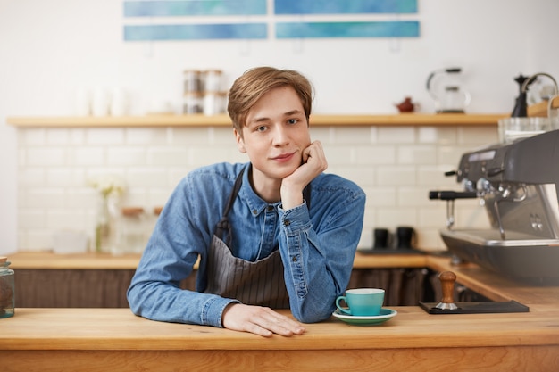 Joli barista masculin portant une chemise en jean et un tablier à rayures grises.