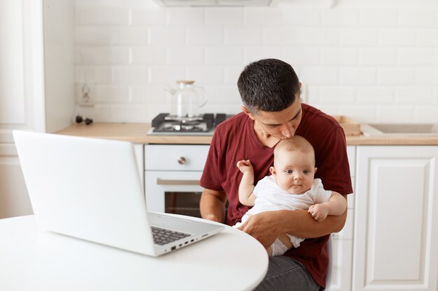 Joli bel homme aux cheveux noirs portant un t-shirt décontracté bordeaux, embrassant sa charmante fille, travaillant sur un ordinateur portable tout en faisant du babysitting, posant dans une cuisine blanche.