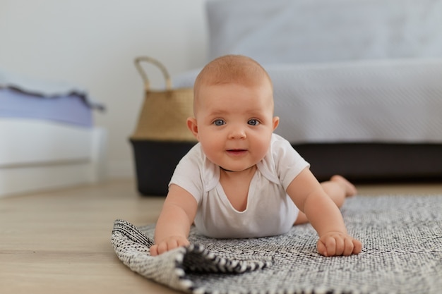 Joli charmant petit bébé garçon ou fille allongé sur le sol essayant de ramper, portant des vêtements blancs, regardant la caméra, s'amusant seul à l'intérieur, enfance heureuse.