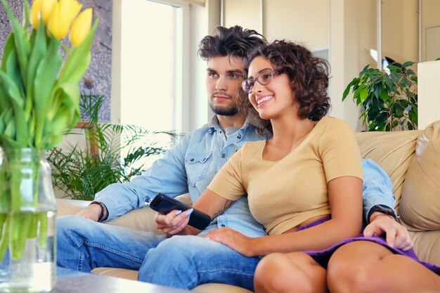 Le joli couple décontracté est assis sur un canapé et change de chaîne de télévision avec une télécommande.