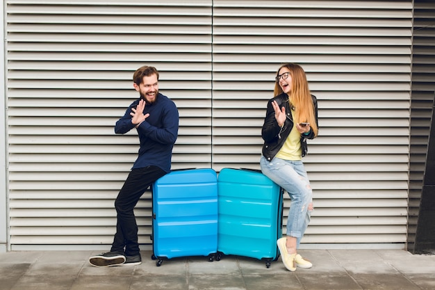 Joli couple est appuyé sur deux valises sur fond rayé gris. Elle a les cheveux longs, des lunettes, un pull jaune, une veste, un jean. Il porte une chemise noire, un pantalon, une barbe. Ils se gratifient mutuellement.