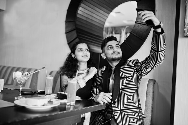 Joli couple indien amoureux portant un sari et un costume élégant assis au restaurant et faisant un selfie par téléphone ensemble Sur la table dessert gâteaux crème glacée et jus