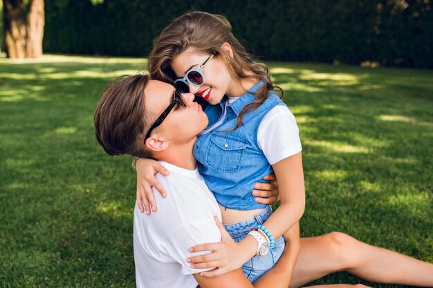 Joli couple de jeunes sur l'herbe dans le parc. Jolie fille aux longs cheveux bouclés en jeans est assise sur un beau mec en T-shirt blanc. Il essaie d'obtenir un baiser.