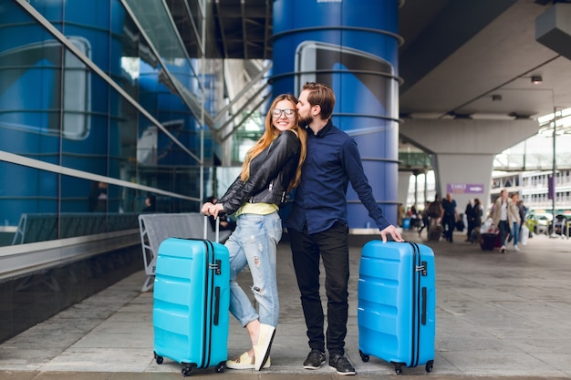 Joli couple avec des valises est debout à l'extérieur de l'aéroport. Elle a les cheveux longs, des lunettes, un pull jaune, une veste. Il porte une chemise noire, une barbe. Guy étreint et embrasse la fille.