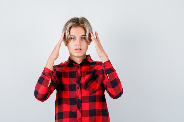 Joli garçon adolescent en chemise à carreaux avec les mains sur la tête et l'air en détresse, vue de face.