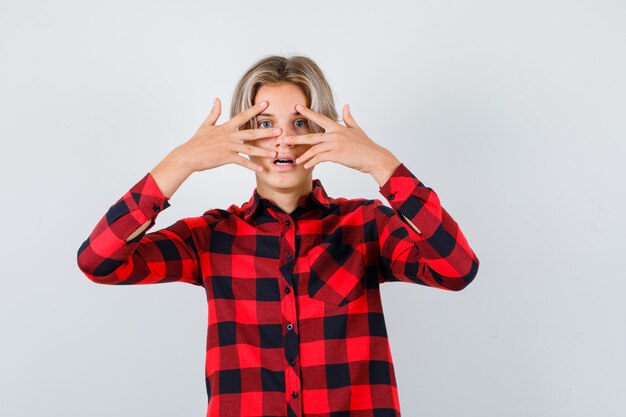 Photo gratuite joli garçon adolescent en chemise à carreaux regardant à travers les doigts et l'air terrifié, vue de face.