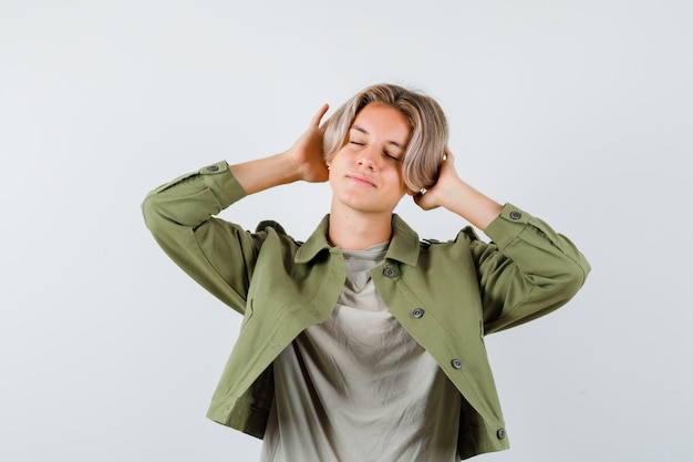 Joli garçon adolescent gardant les mains derrière les oreilles, fermant les yeux en veste verte et l'air ravi, vue de face.