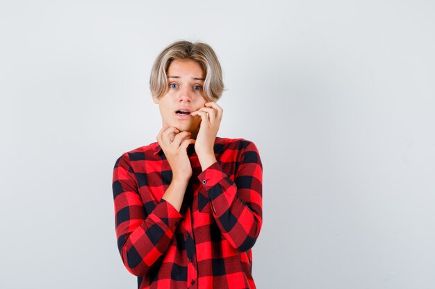 Joli garçon adolescent avec la main sur la joue en chemise à carreaux et l'air terrifié, vue de face.