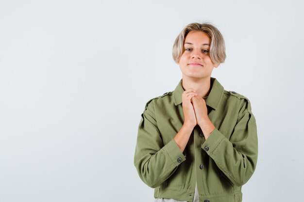 Joli garçon adolescent serrant les mains dans un geste de prière en veste verte et à la recherche de joie. vue de face.