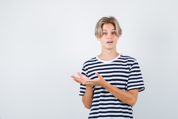 Joli garçon adolescent en t-shirt rayé faisant semblant de montrer quelque chose et ayant l'air perplexe, vue de face.