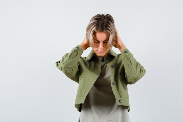 Joli garçon adolescent en veste verte gardant les mains sur les oreilles et ayant l'air en détresse, vue de face.