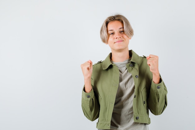 Joli garçon adolescent en veste verte montrant le geste du gagnant et l'air joyeux, vue de face.