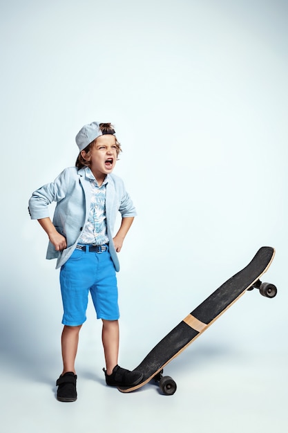 Joli jeune garçon sur planche à roulettes dans des vêtements décontractés sur le mur blanc du studio