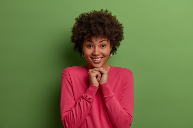 Photo gratuite joli jeune modèle féminin aux cheveux bouclés à la peau sombre garde les mains pressées ensemble sous le menton, sourit doucement, montre des dents blanches parfaites, porte des vêtements roses, des modèles contre le mur vert