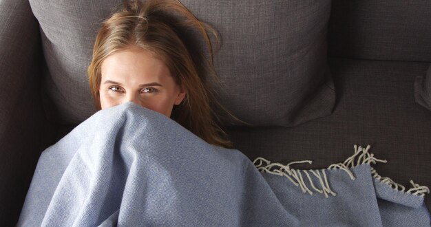 Joli modèle féminin jouant au coucou avec une fine couverture bleu clair allongée sur un canapé gris foncé