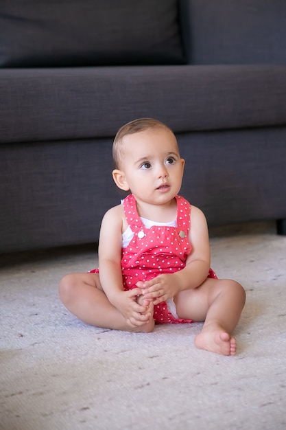 Photo gratuite joli petit bébé assis sur un tapis pieds nus dans le salon. jolie fille réfléchie en short salopette rouge regardant quelqu'un et touchant le pied. concept de week-end, enfance et être à la maison