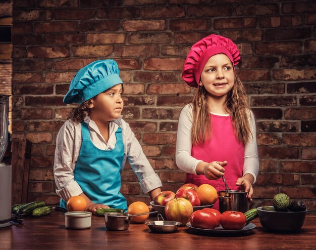 Joli petit couple de cuisiniers. Petit garçon aux cheveux bouclés bruns vêtu d'un uniforme de cuisinier bleu et une belle fille vêtue d'un uniforme de cuisinier rose cuisinant ensemble dans une cuisine contre un mur de briques.