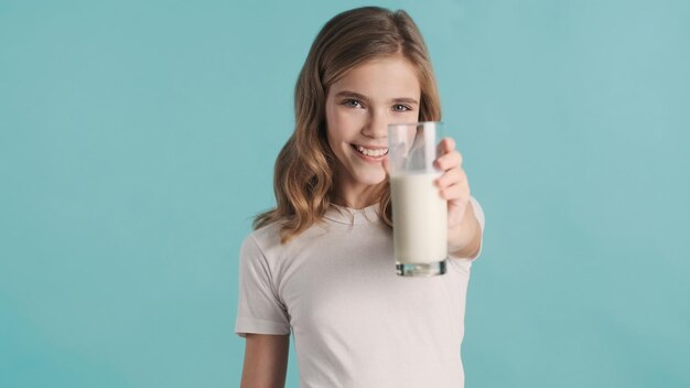 Jolie adolescente blonde souriante aux cheveux ondulés offrant un verre de lait à la caméra sur fond bleu