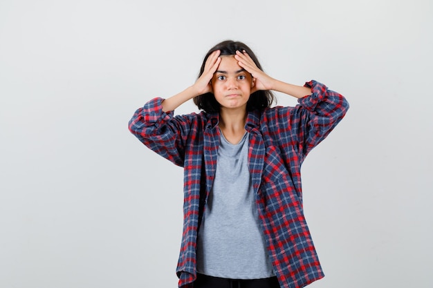 Jolie adolescente en chemise à carreaux tenant les mains sur la tête et l'air confus, vue de face.