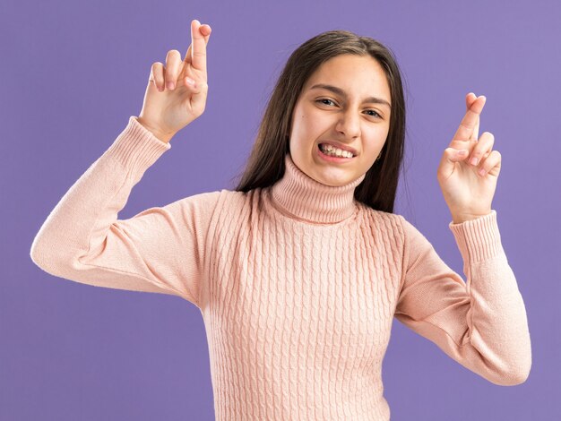 Une jolie adolescente pleine d'espoir faisant un geste de bonne chance avec les deux mains isolées sur un mur violet