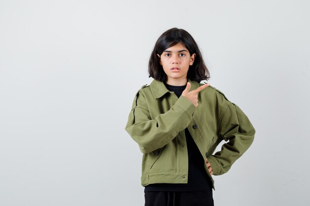 Jolie adolescente pointant vers le coin supérieur droit en veste verte de l'armée et l'air perplexe. vue de face.