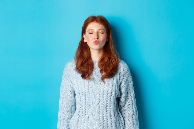 Jolie adolescente rousse en attente de baiser, lèvres plissées et yeux fermés, debout en pull sur fond bleu.