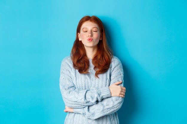 Jolie adolescente rousse en attente de baiser, lèvres plissées et yeux fermés, debout en pull sur fond bleu.