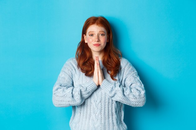 Jolie adolescente rousse demandant de l'aide, souriante en quémandant une faveur, a besoin de quelque chose, debout sur fond bleu