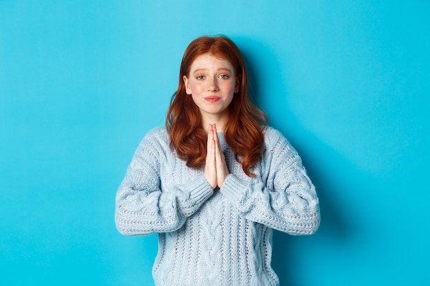 Photo gratuite jolie adolescente rousse demandant de l'aide, souriante en quémandant une faveur, a besoin de quelque chose, debout sur fond bleu