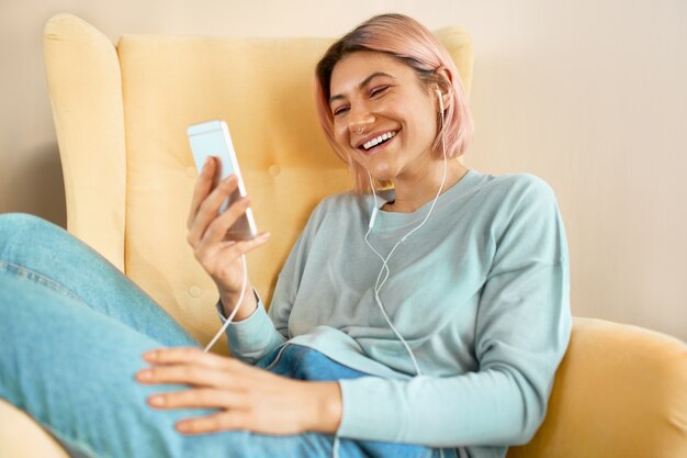 Jolie adolescente se détendre à la maison en discutant avec des amis en ligne sur mobile via un appel webcam de groupe, souriant. Jolie jeune femme assise dans un fauteuil avec téléphone portable et écouteurs, regarder une vidéo drôle