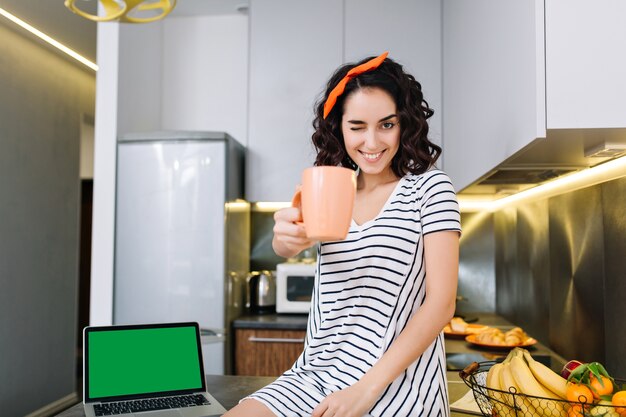 Jolie belle jeune femme aux cheveux bouclés coupés souriant avec une tasse de thé dans la cuisine dans un appartement moderne. Bonjour, confort à la maison, week-end, s'amuser