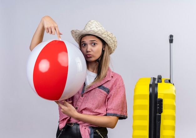 Une jolie belle jeune femme portant une chemise rouge et un chapeau tenant ballon gonflable sur un mur blanc
