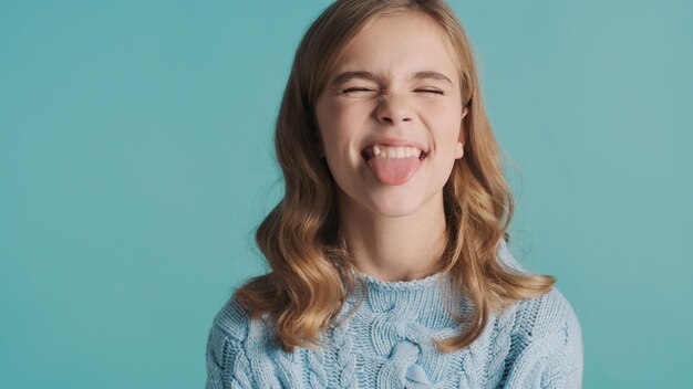 Jolie blonde adolescente aux cheveux ondulés montrant la langue sur l'appareil photo à la drôle sur fond bleu