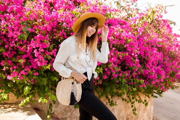 Jolie blonde femme au chapeau de paille posant sur un arbre en fleurs roses au printemps ensoleillé