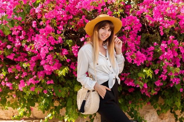 Jolie blonde femme au chapeau de paille posant sur un arbre en fleurs roses au printemps ensoleillé