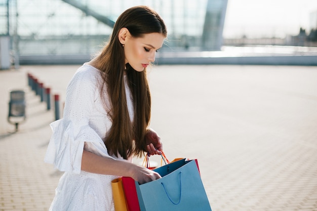 Jolie brune aux cheveux longs regarde à l&#39;intérieur des sacs à provisions devant un bâtiment moderne