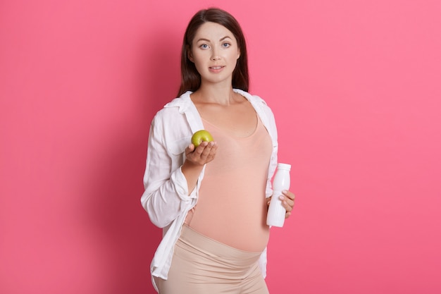 Jolie brune enceinte tenant une pomme verte et une bouteille de lait ou de yaourt, regarde la caméra, porte une chemise et des leggins, femme enceinte, manger des aliments sains.