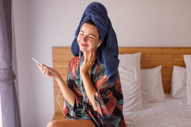 Jolie brune femme enveloppée dans une serviette de douche à l'aide d'un téléphone portable assis sur le lit