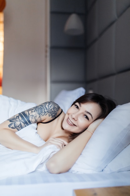 Jolie brune femme avec tatouage se pose dans le lit sur l'oreiller le matin dans l'appartement de mode