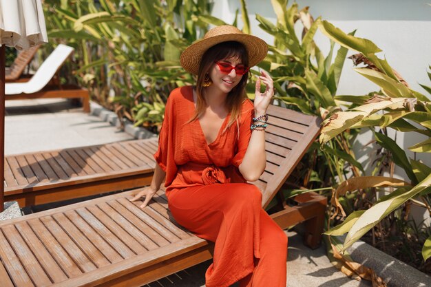 Jolie brune femme en tenue orange élégante et chapeau de paille au froid sur une chaise longue près de la piscine.