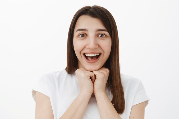 Jolie brune impressionnée et accablée en t-shirt blanc écoutant attentivement l'événement étonnant souriant ravi expremely amusé se tenant la main au-dessus de la mâchoire comme hypnotisé et stupéfait par des nouvelles impressionnantes