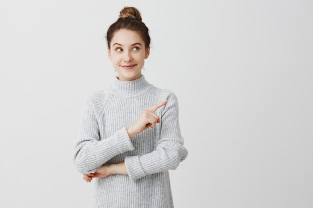 Jolie brunette femme posant avec un regard délicat et gesticulant sur le côté. Designer féminin présentant un joli produit avec l'index. Concept de geste