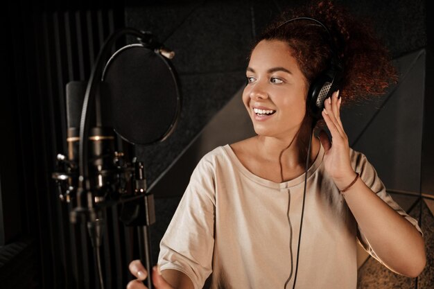 Jolie chanteuse écoutant du son dans des écouteurs enregistrant joyeusement une nouvelle chanson dans un studio d'enregistrement professionnel