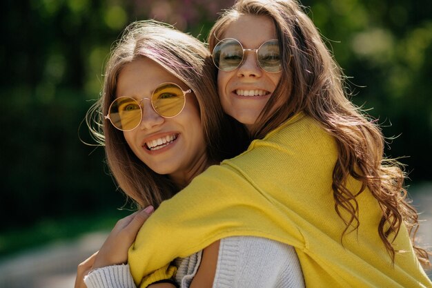 Jolie charmante dame aux longs cheveux ondulés en pull brillant étreint sa sœur et rit Deux filles élégantes souriant au soleil et portant des lunettes de soleil rondes et des pulls brillants