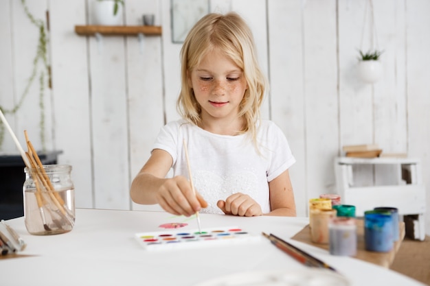 Jolie et charmante fille blonde portant un t'shirt blanc approfondissant la brosse dans la peinture