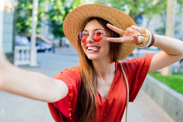 Jolie dame au chapeau élégant faisant selfie tout en marchant en plein air les week-ends d'été
