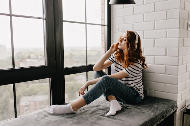 Photo gratuite jolie dame au gingembre en jeans assis sur le rebord et écoute de la musique. heureux fille bouclée posant à côté de la fenêtre dans les écouteurs.